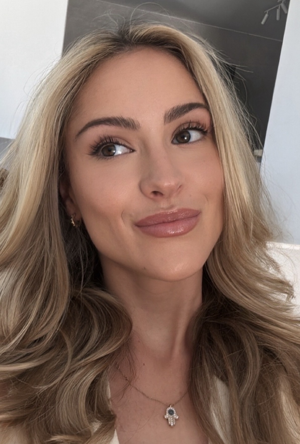 Close-up of a smiling woman with long blonde hair wearing a necklace with a hamsa hand pendant.