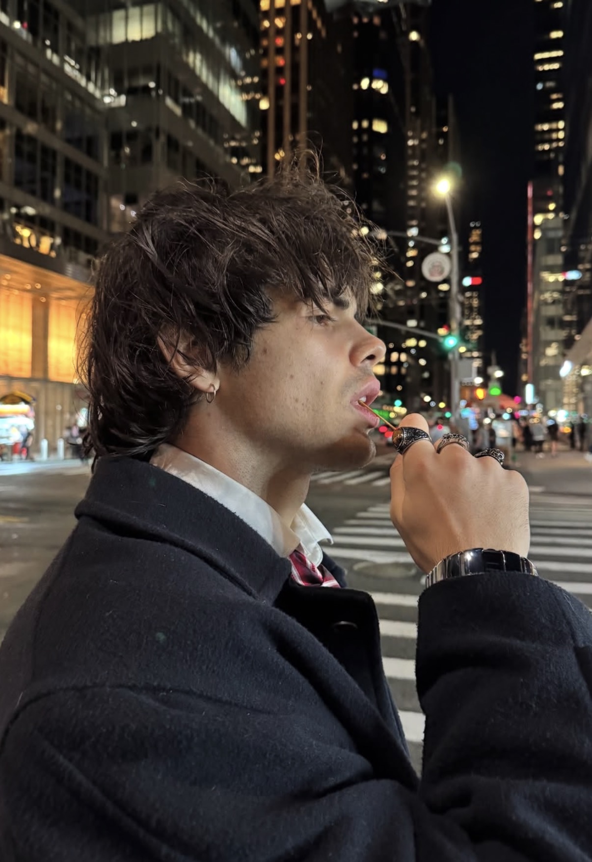 Young man with tousled hair wearing a dark coat and rings, licking a lollipop on a city street at night.