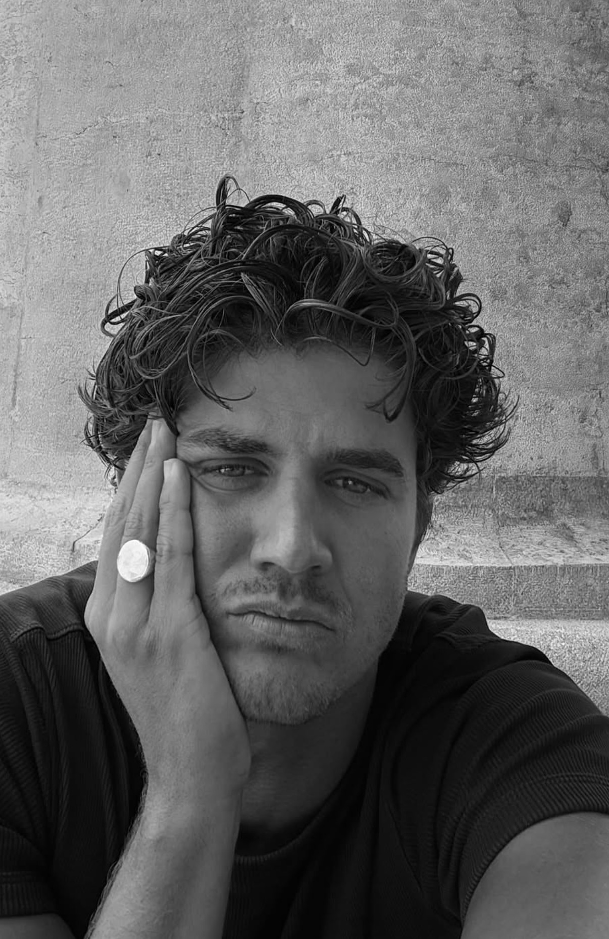 Black and white close-up of a man with curly hair resting his face on his hand, wearing a large ring and a dark shirt.