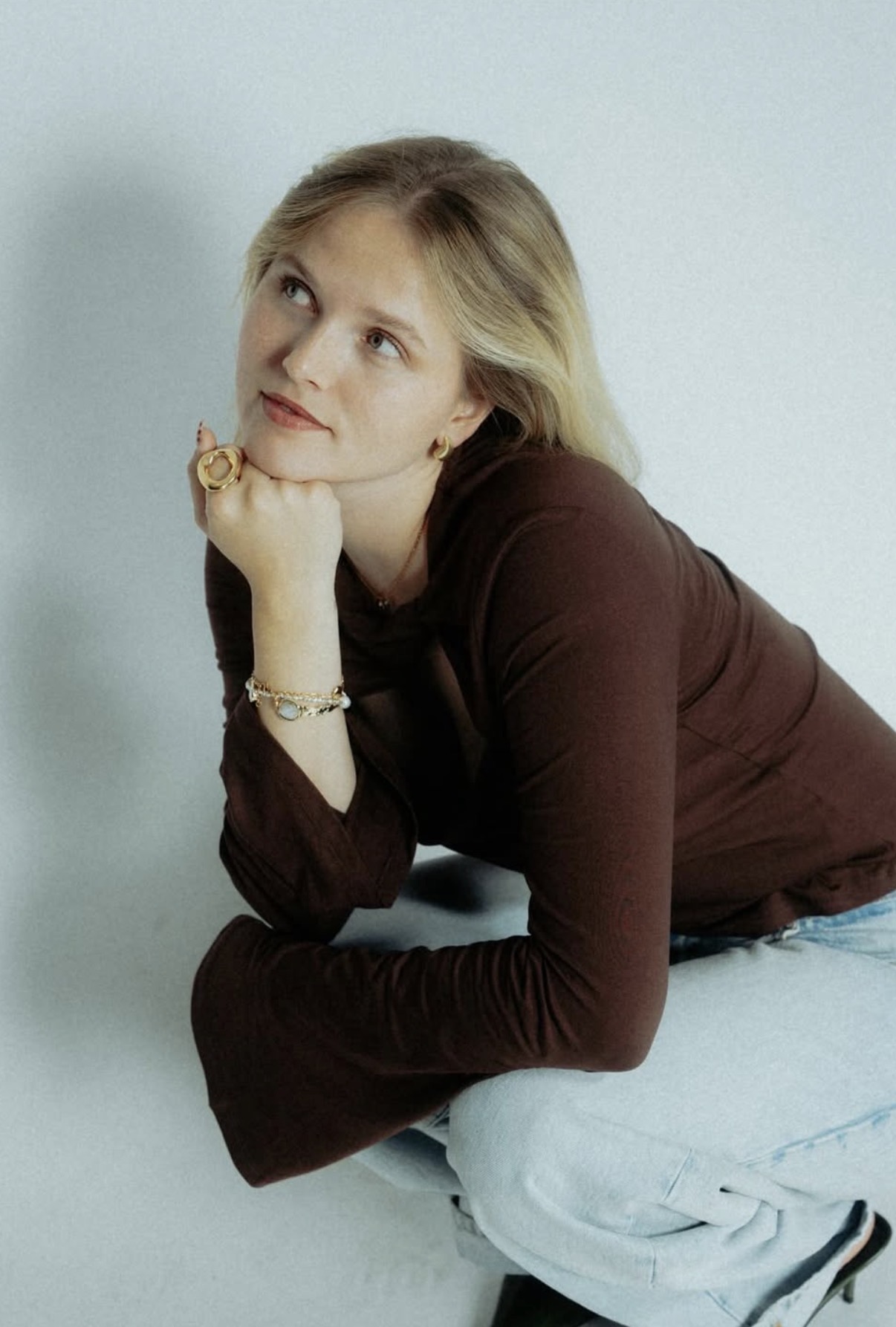 Blonde woman in a dark brown long-sleeve top and light blue jeans, sitting with chin resting on hand and looking slightly upward.
