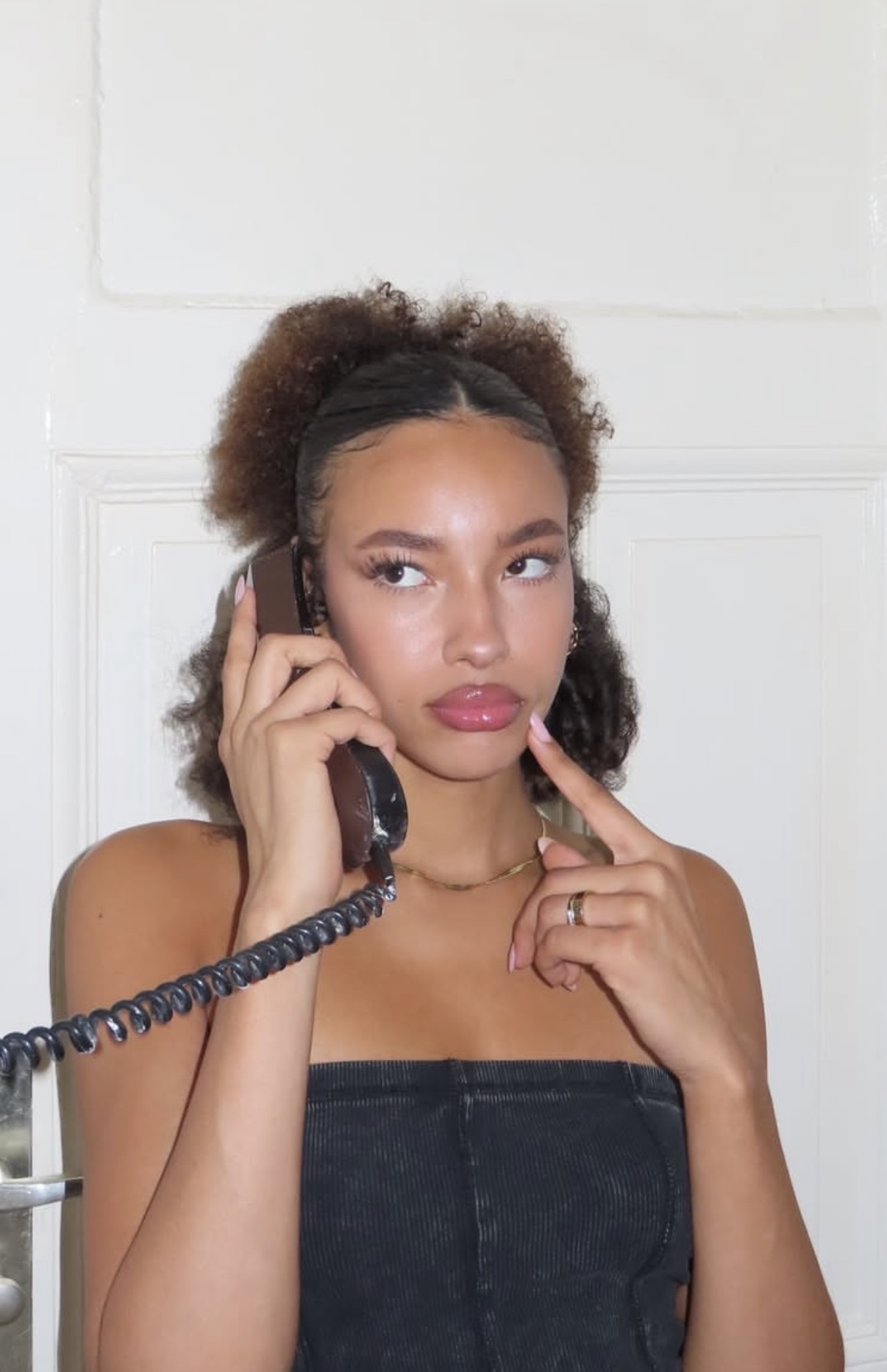 Young woman with curly hair holding a corded phone to her ear, looking thoughtfully to the side with a finger touching her chin.