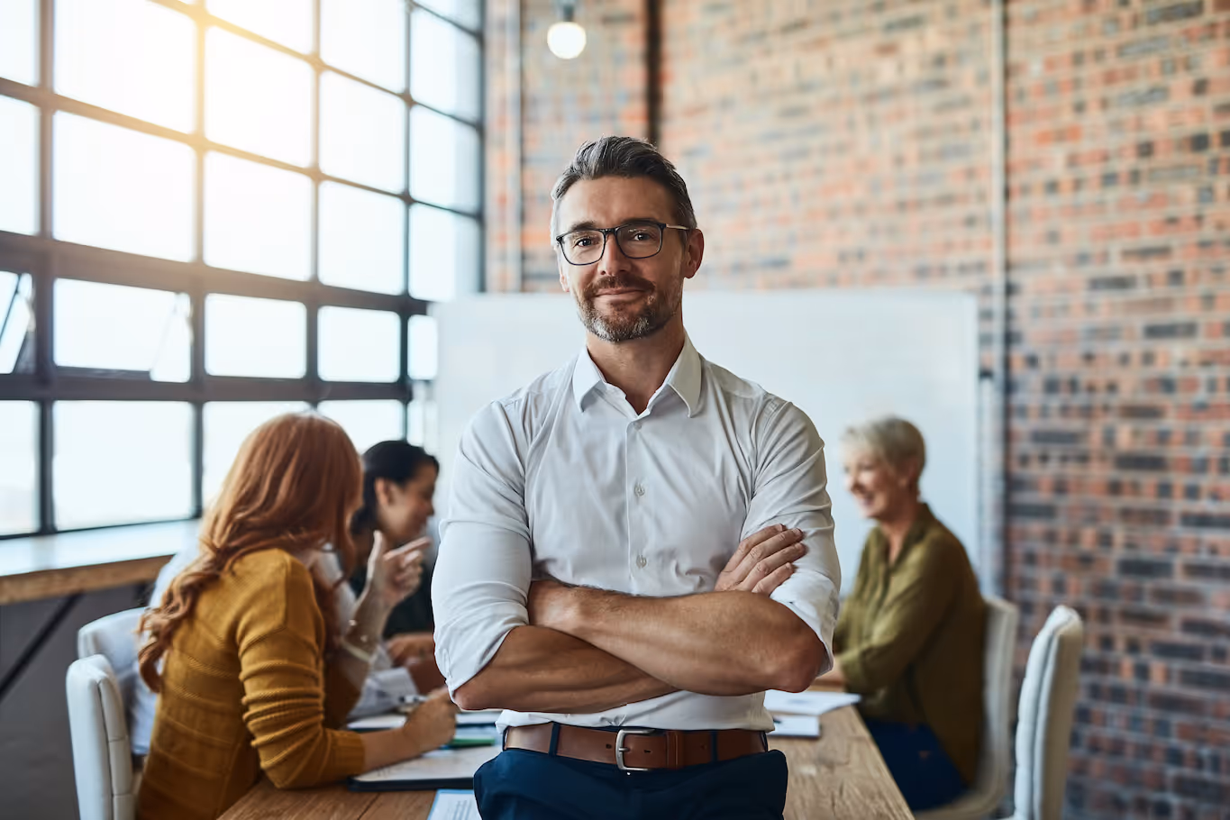 zufriedener Unternehmer lächelt, Arme verschränkt, confident and happy business man in an office with working employees behind him