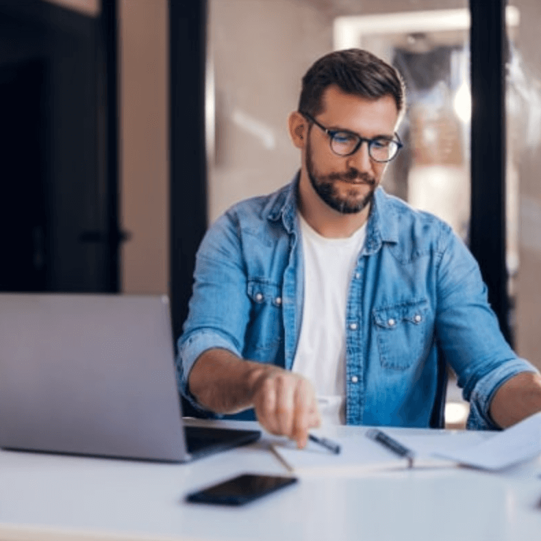 Geschäftsmann mit Brille arbeitet konzentriert am Schreibtisch vor einem Laptop und überprüft Dokumente – Symbolbild für Finanzierungsantrag.