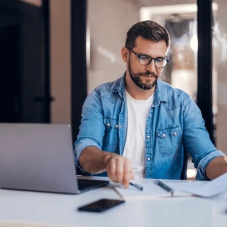 Geschäftsmann mit Brille arbeitet konzentriert am Schreibtisch vor einem Laptop und überprüft Dokumente – Symbolbild für Finanzierungsantrag.