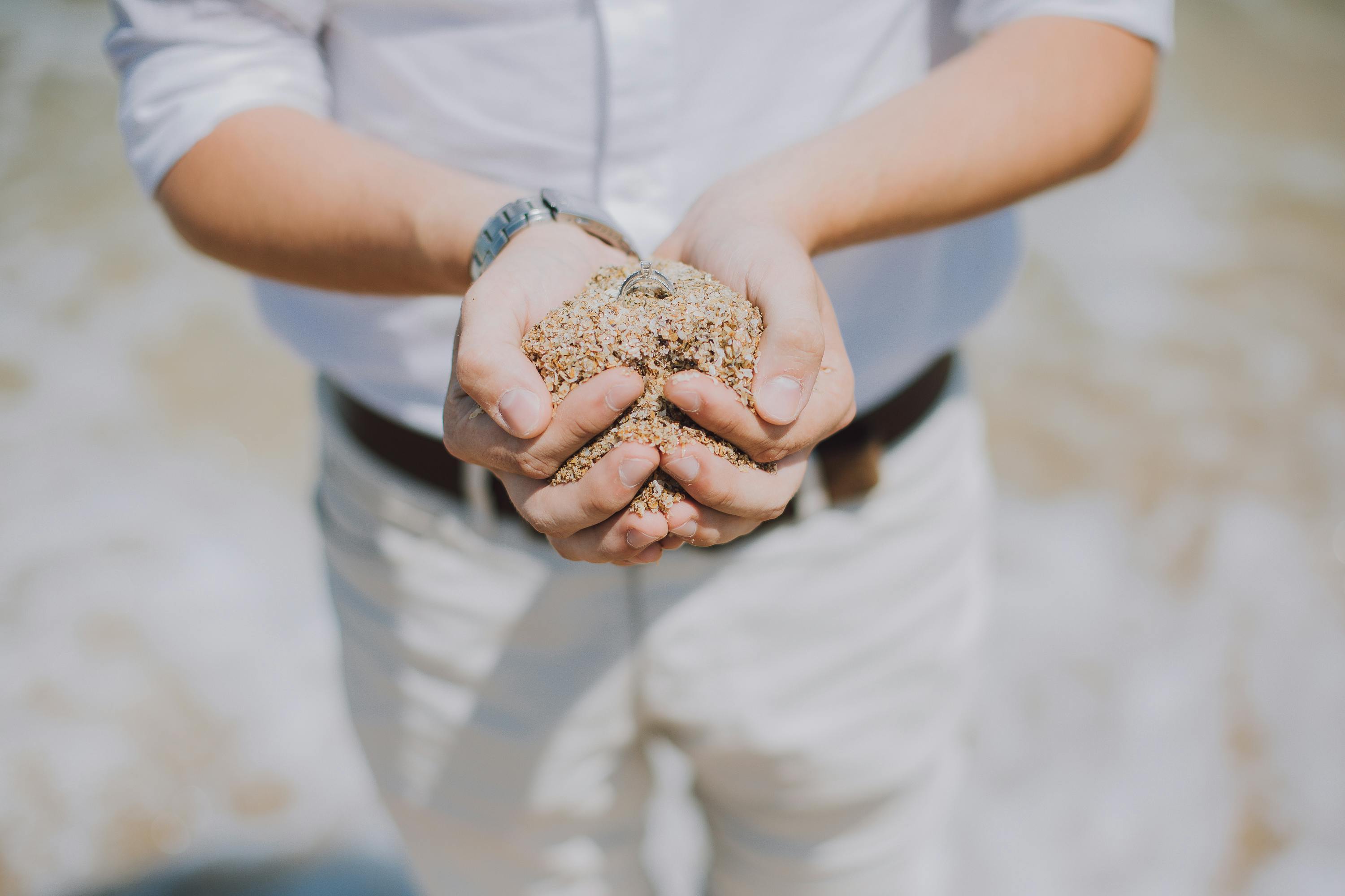 Person holding a handful of sand with a silver ring partially buried in the sand.