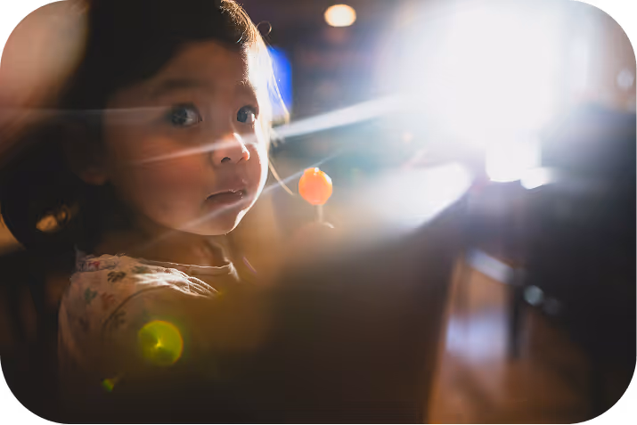 Little girl reading a large illustrated book in a dimly lit room, with warm light and holiday decorations behind her.