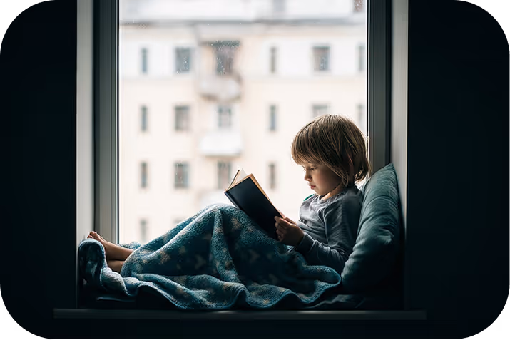 Little girl reading a large illustrated book in a dimly lit room, with warm light and holiday decorations behind her.