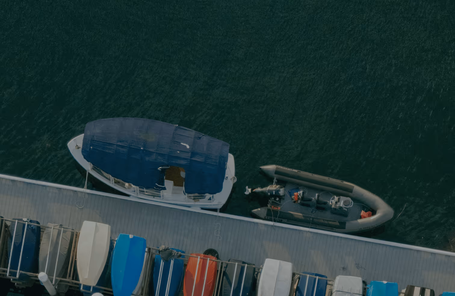 Aerial view of two boats docked alongside a wooden pier with several covered kayaks or small boats lined up on the pier.