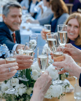 Group of people clinking glasses of champagne at a celebratory gathering with white flowers on the table.