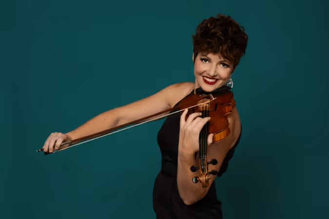 Smiling woman with short curly hair wearing a black sleeveless dress playing a violin against a teal background.