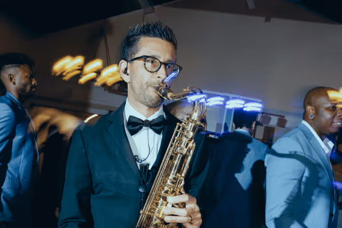 Man in black tuxedo playing a saxophone at a party or event with people in suits in the background.