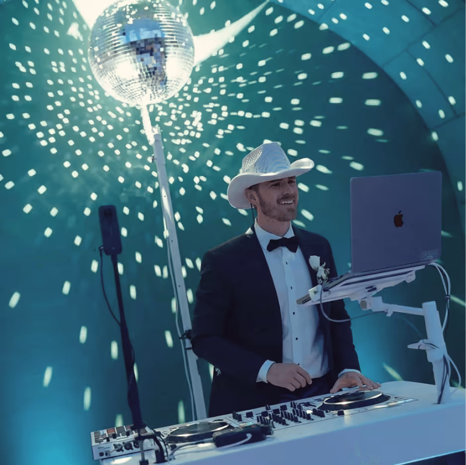 Man in tuxedo and white cowboy hat DJing with a laptop and mixer under a disco ball casting light spots.