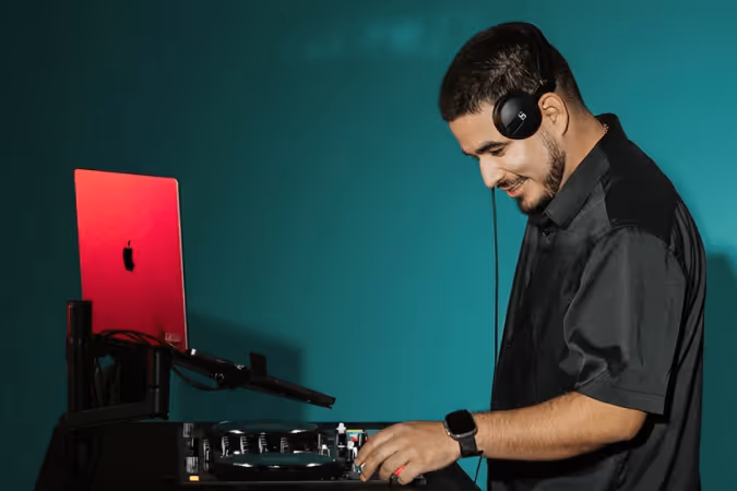 DJ wearing headphones and a black shirt using a turntable and laptop against a teal background.