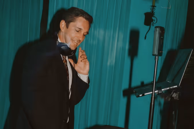 Man in a suit wearing headphones and smiling while standing near a microphone and laptop in front of teal curtains.