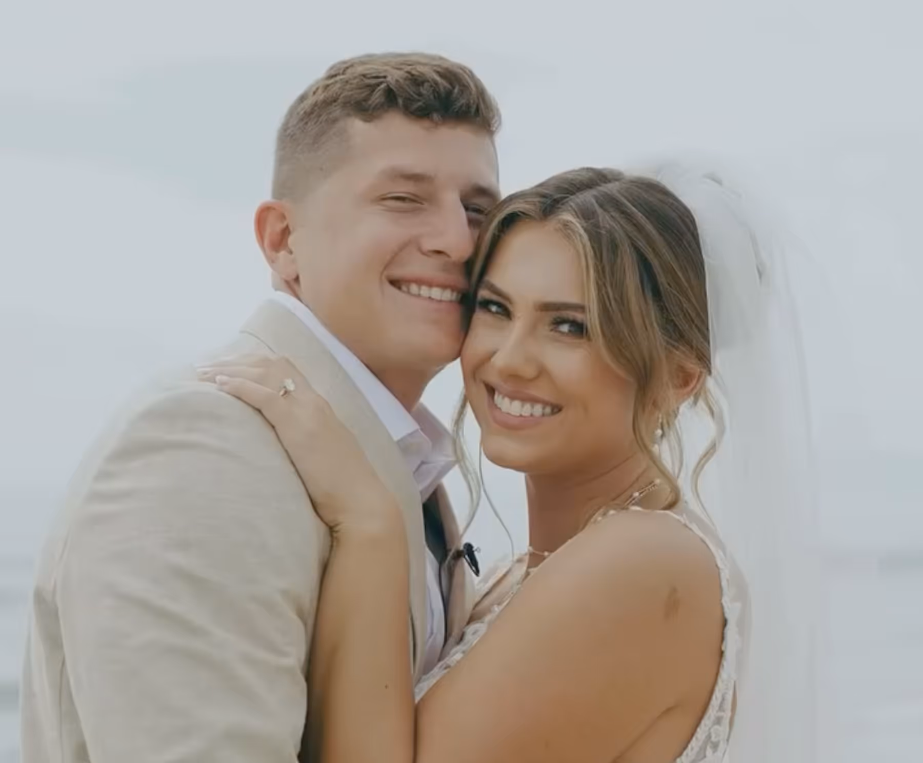Smiling bride in veil embracing groom in light-colored suit outdoors.