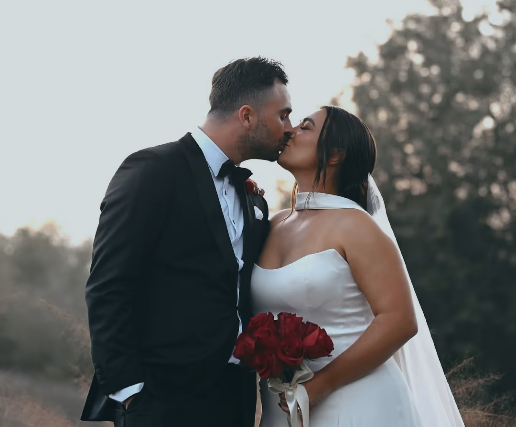 Bride and groom in wedding attire sharing a kiss outdoors with bride holding a bouquet of red roses.