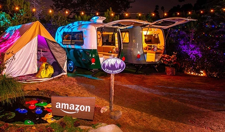 Outdoor nighttime scene with a tent and two travel trailers, illuminated by string lights, featuring Amazon and Camp Ground signs