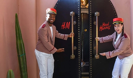 Two hotel staff members with blurred faces opening double doors labeled ‘H&M’ and ‘Hotel Hennes’
