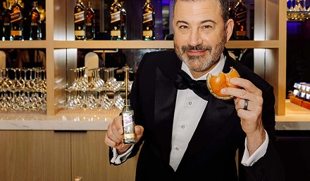 Man in a tuxedo holding a cocktail and a bottle, face intentionally blurred for privacy, standing in front of a bar