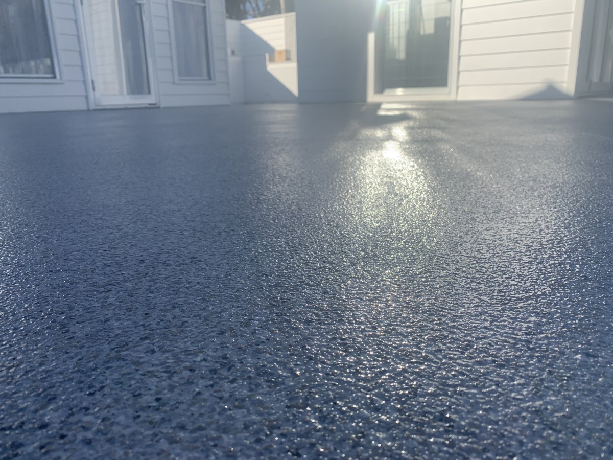 Close-up of a textured, glistening dark gray patio floor with sunlight reflecting off its surface and white siding walls with doors in the background.