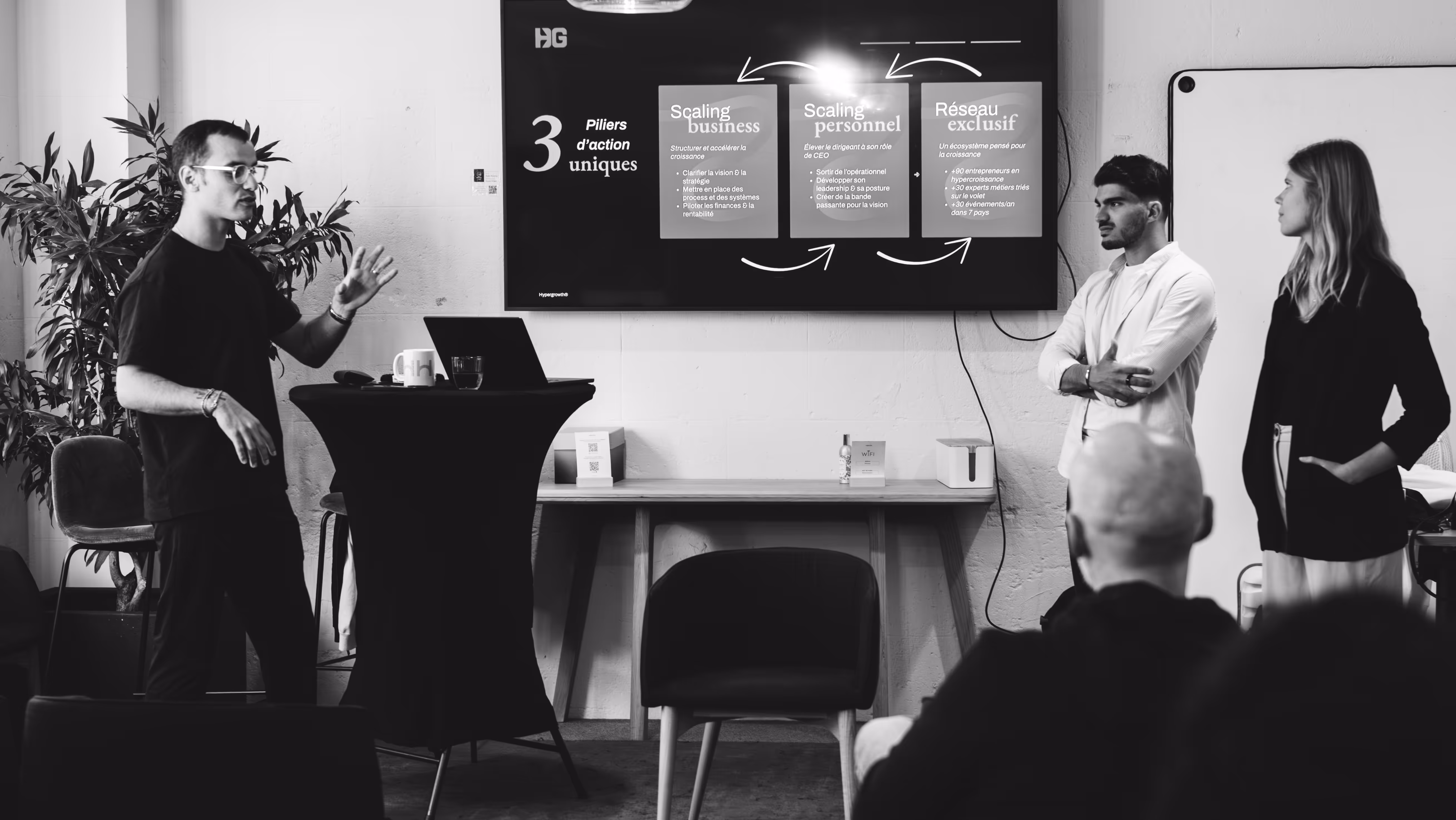 3 operating partners debout sur une estrade devant une présentation proposant des solutions business aux entrepreneurs présents dans la pièce lors d'une session de networking.