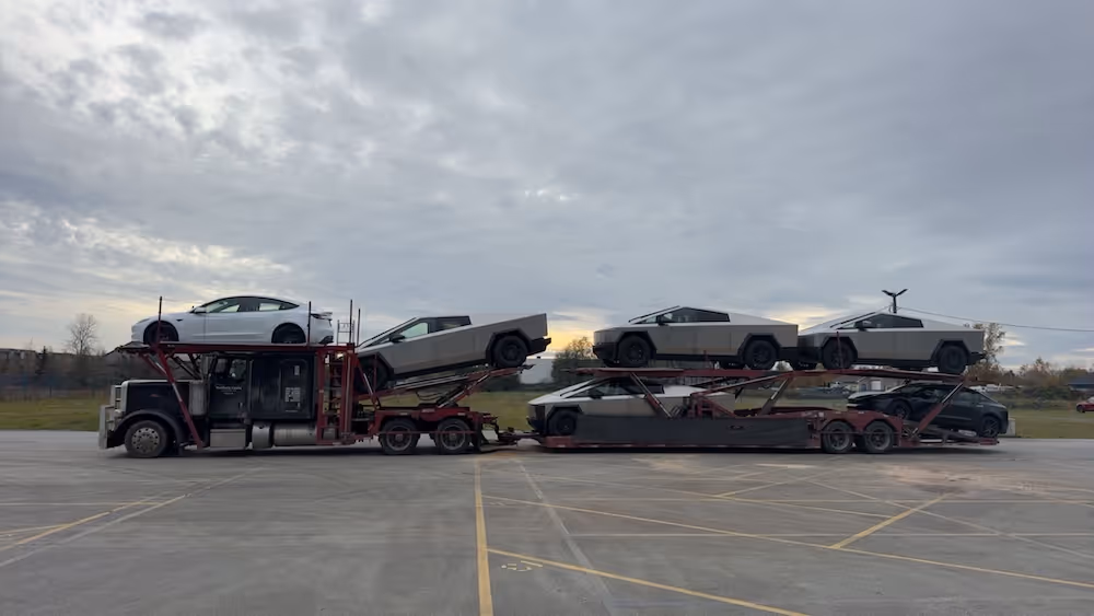 Car carrier truck loaded with one white Tesla Model Y and four gray Tesla Cybertrucks parked on an empty paved lot under a cloudy sky.