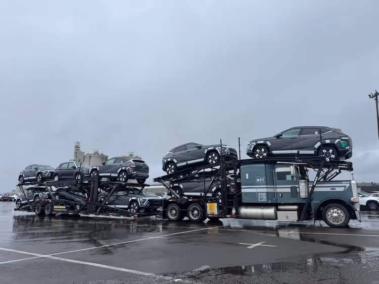 Gray car hauler truck loaded with two levels of dark gray SUVs parked on wet pavement under an overcast sky.