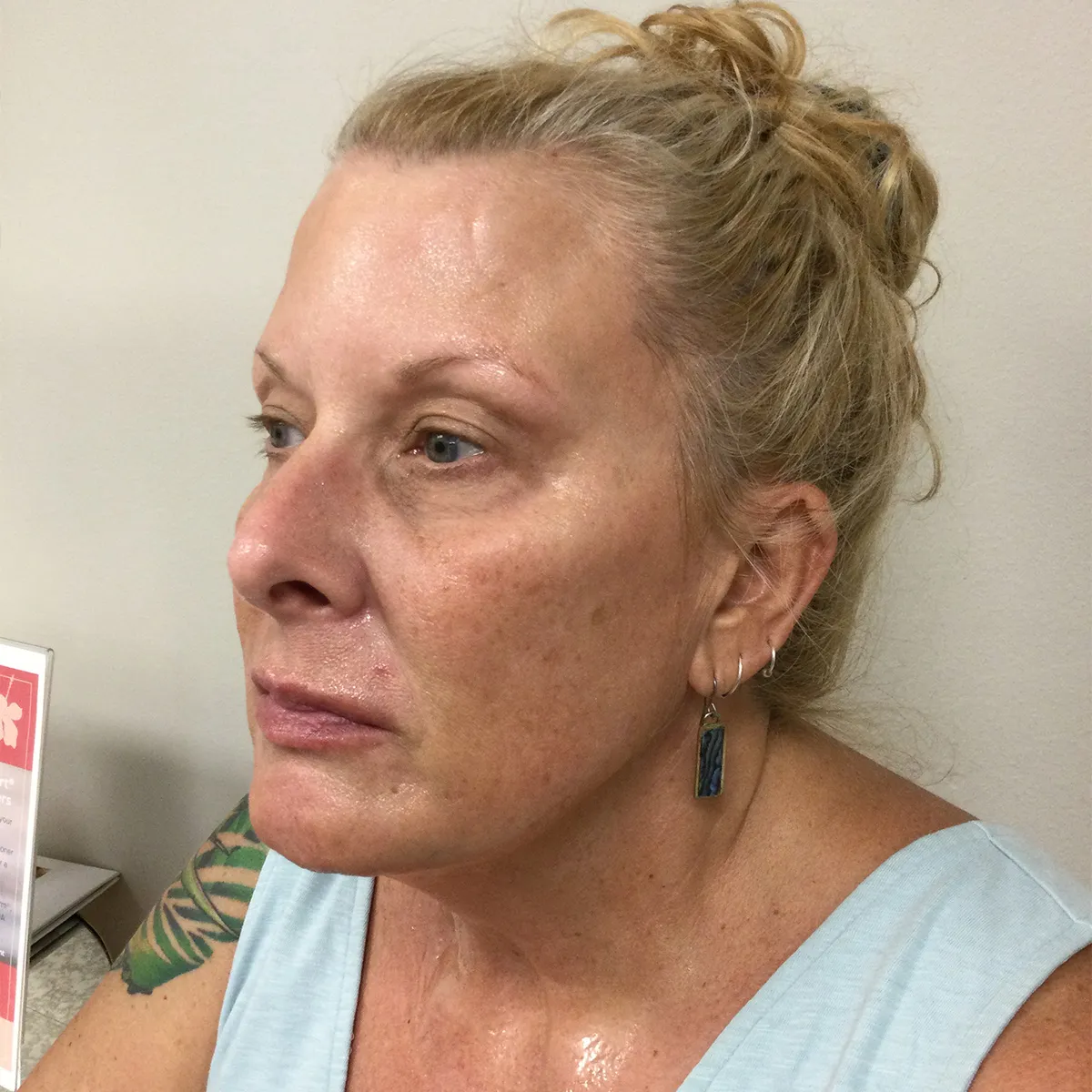 Close-up profile of a middle-aged woman with light hair in a bun, wearing a light blue sleeveless top and a rectangular earring, against a plain background.