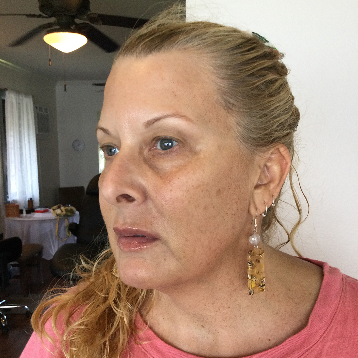 Close-up of a woman with blonde hair tied back wearing dangly earrings and a pink top indoors.