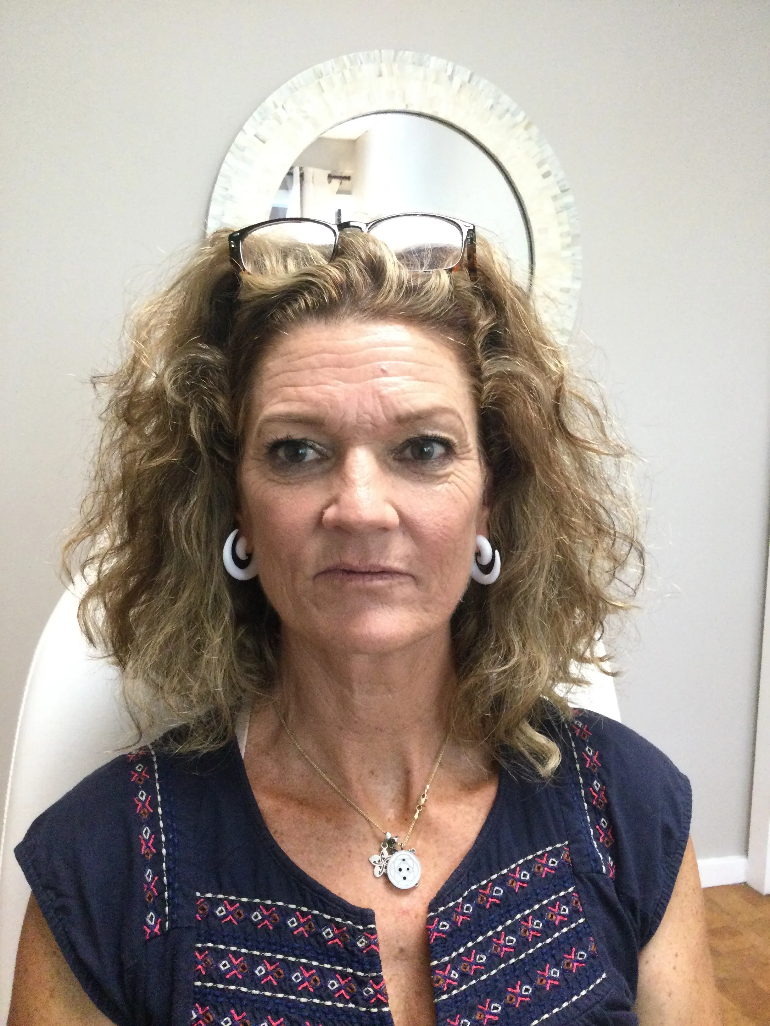 Middle-aged woman with curly blonde hair wearing white hoop earrings and a navy blue top with red and white embroidery, with glasses resting on her head and a decorative round pendant necklace.