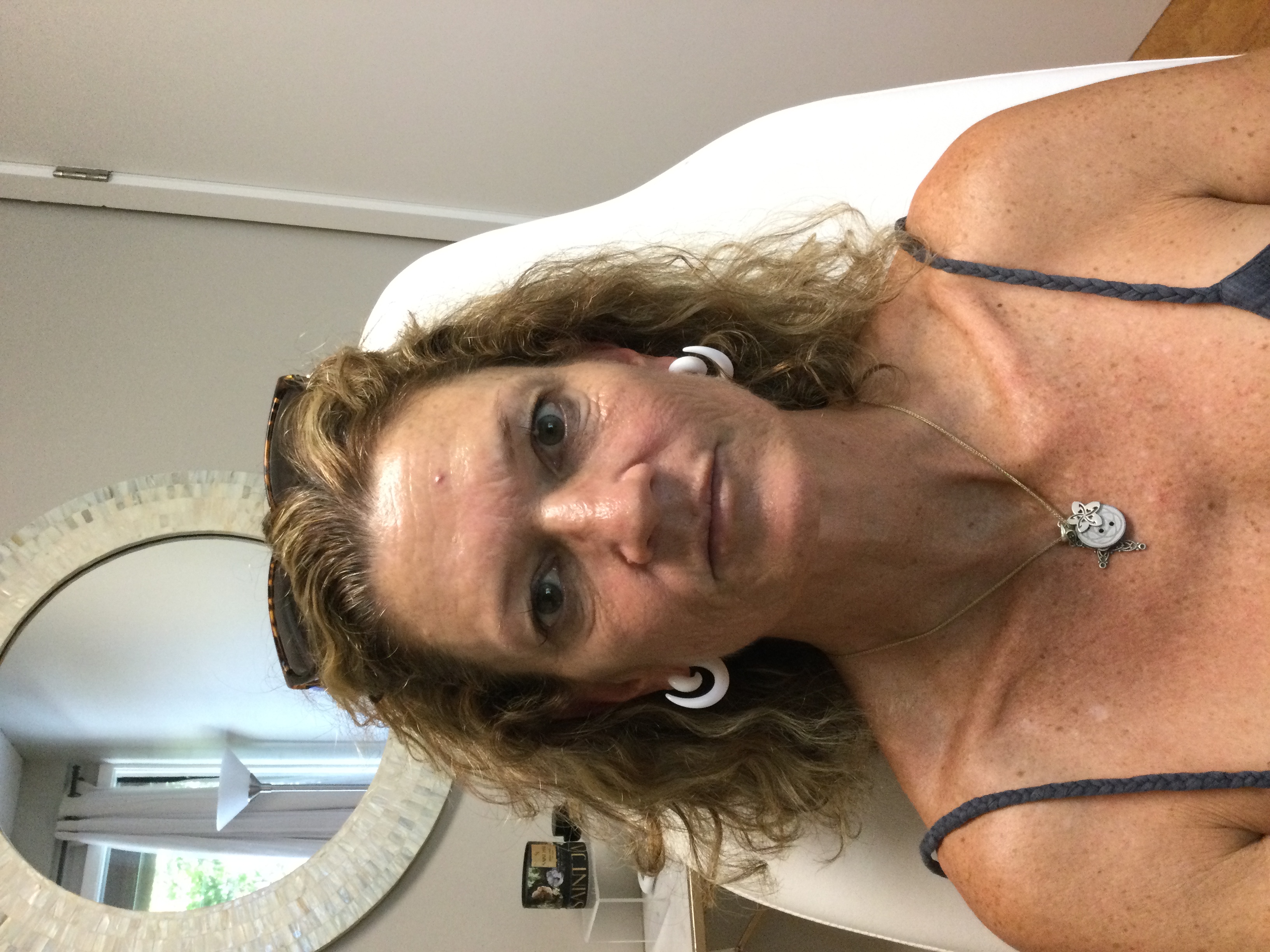 Middle-aged woman with curly hair wearing white hoop earrings, a necklace with multiple pendants, and a dark tank top sitting in a white chair indoors.