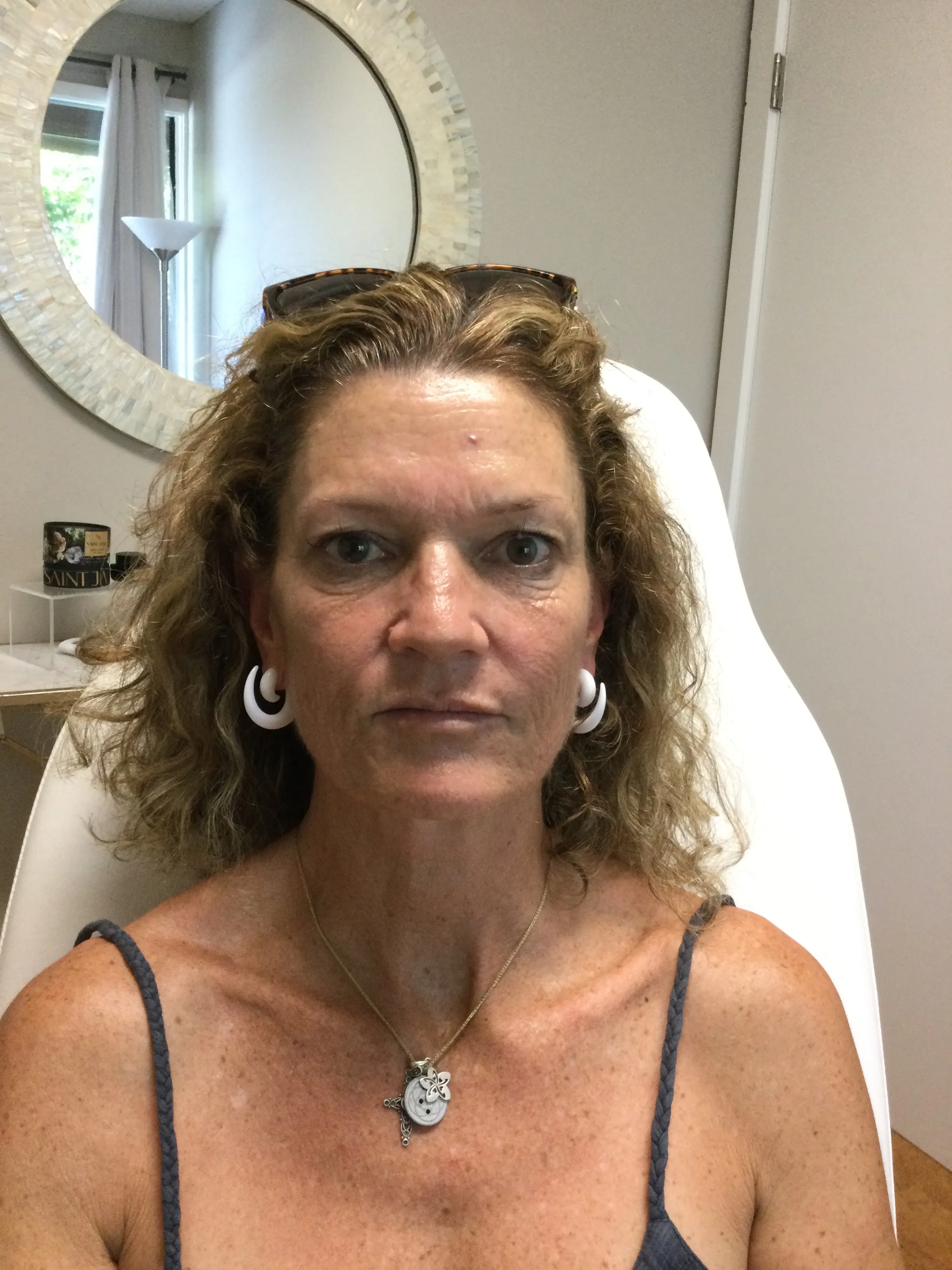 Middle-aged woman with curly hair wearing white hoop earrings, a necklace with multiple pendants, and a dark tank top sitting in a white chair indoors.