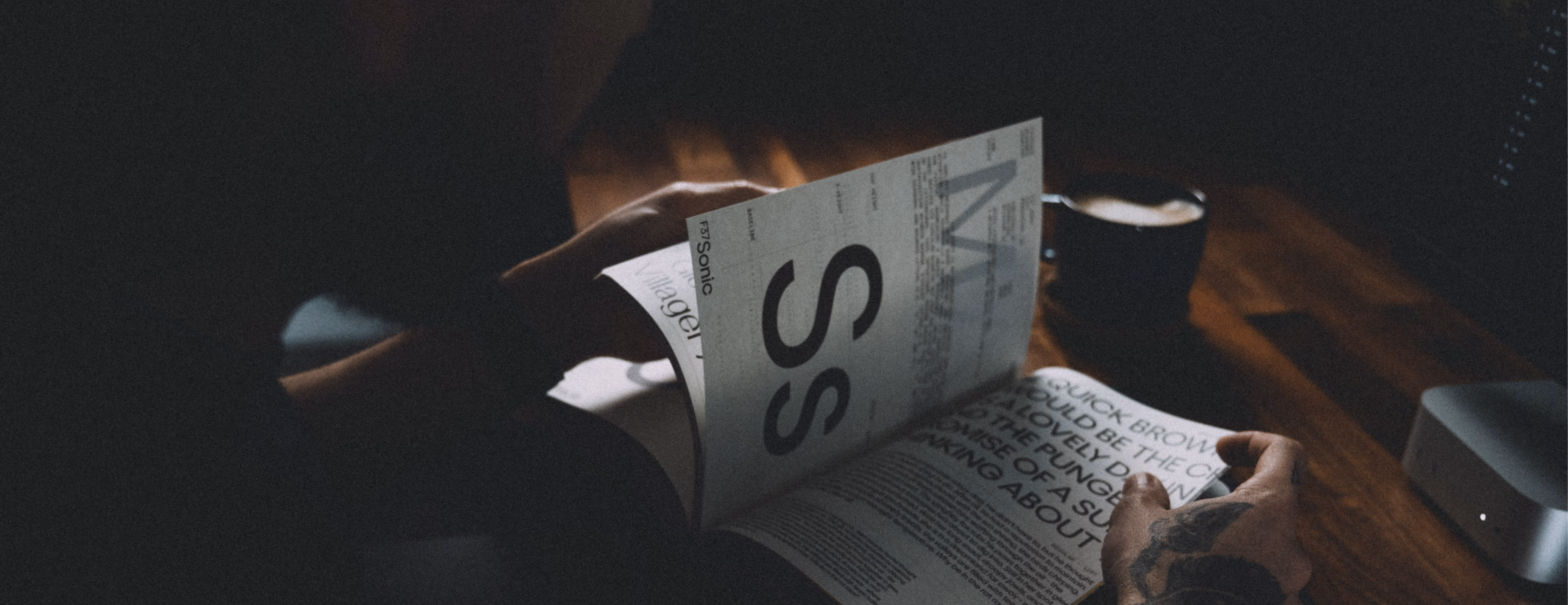 Person with tattooed hand reading a magazine by a wooden desk with a cup of coffee and electronic device.