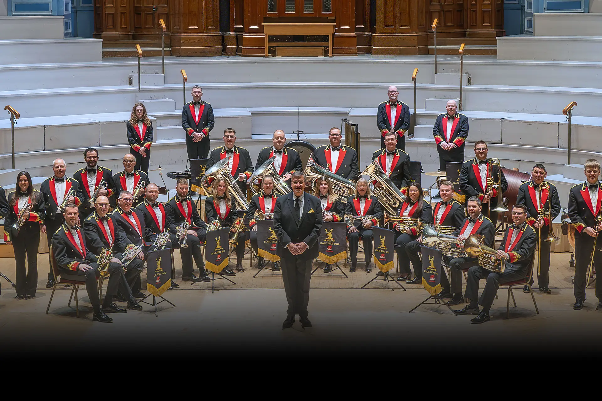 Black Dyke Band in ceremonial red-and-black uniform on stage, Music Director Nicholas Childs at the front.