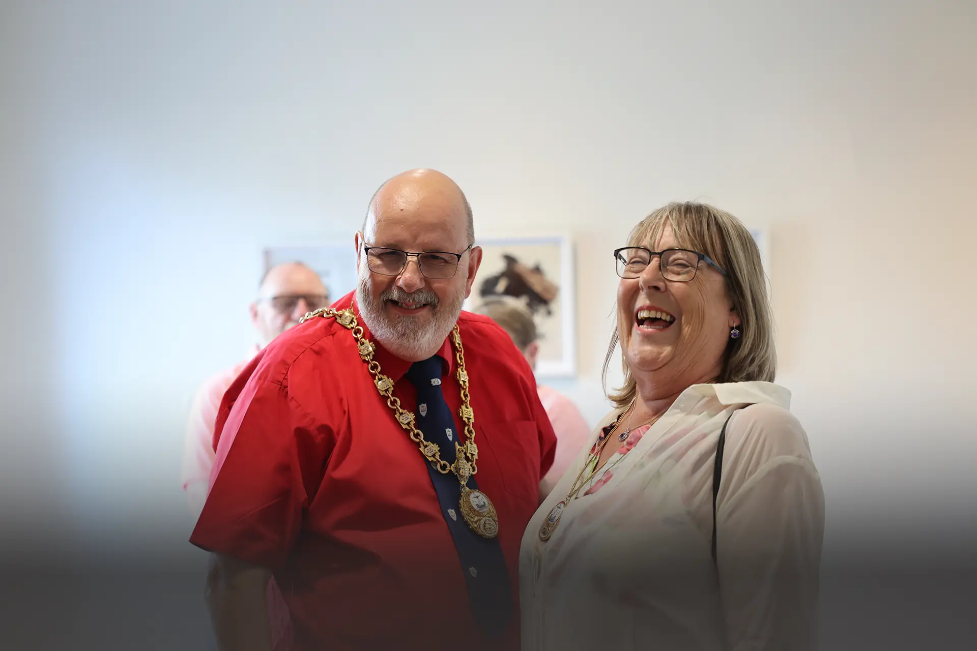 The Mayor and Mayoress of New Romney share a laugh at a JAM private view, framed artwork displayed on the wall behind them.