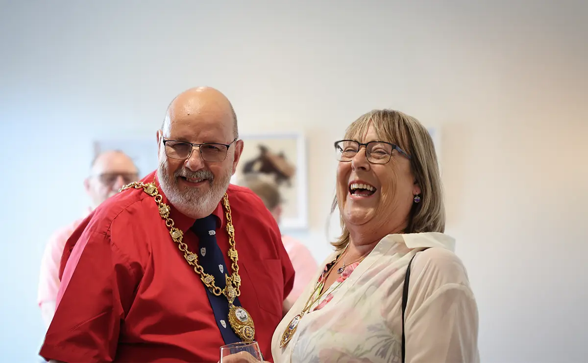 The Mayor and Mayoress of New Romney share a laugh at a JAM private view, framed artwork displayed on the wall behind them.