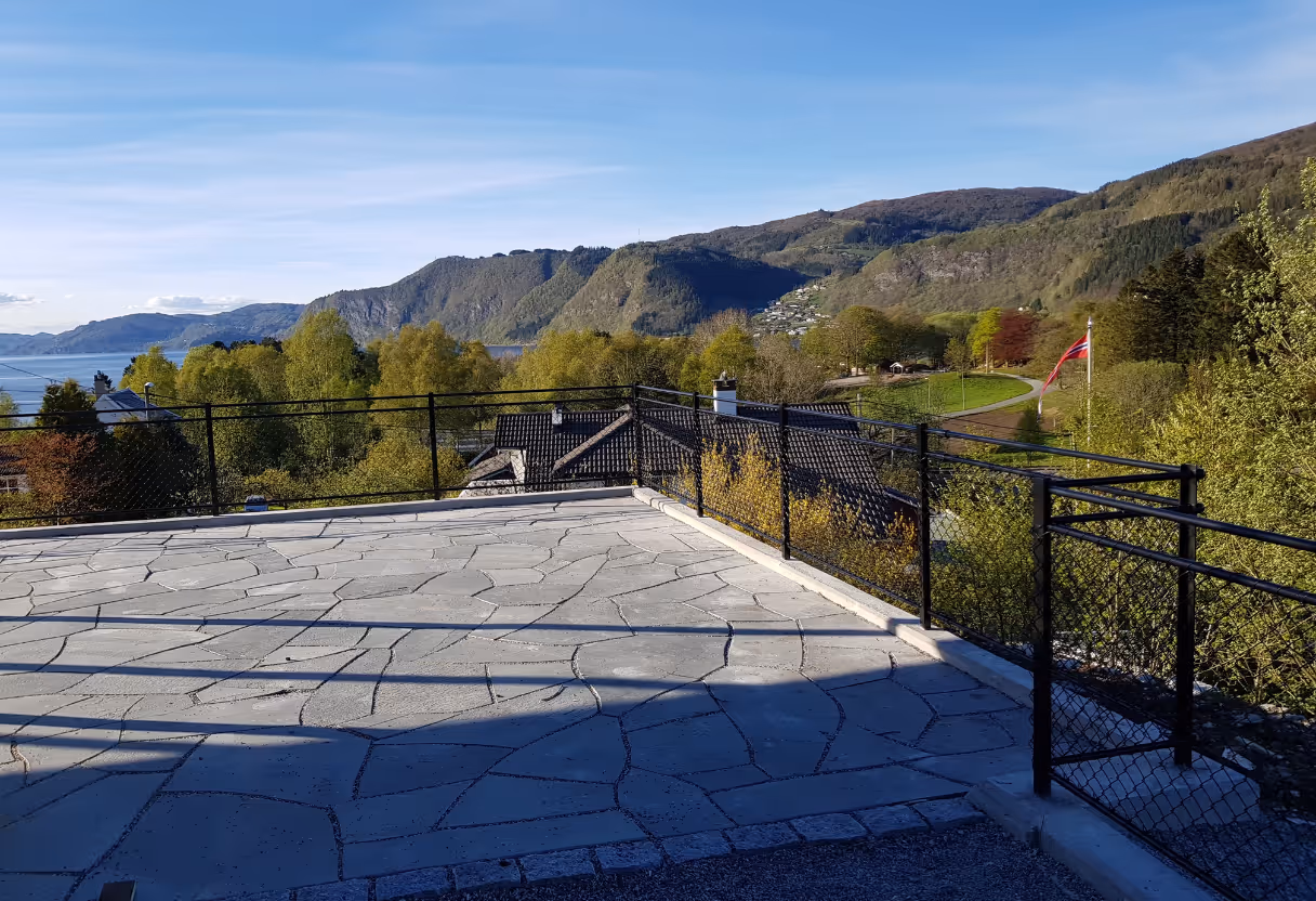 Utsikt fra en steinlagt terrasse med svart rekkverk mot grønne trær, fjell og en norsk flagg på en flaggstang.