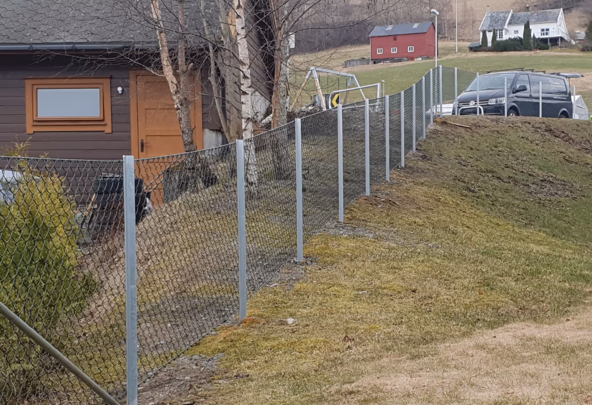 Kjettinglenkegjerde langs en skråning ved et hus og parkerte biler i bakgrunnen.