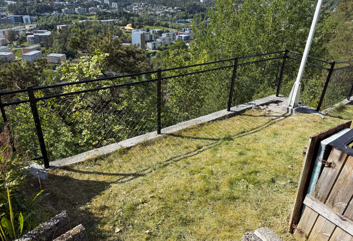 Svart nettinggjerde på en plenen med utsikt over bygninger og skog i bakgrunnen.