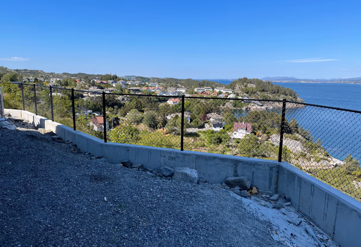 Sikt mot en kystby med et svart nettinggjerde på en betongmur under en blå himmel.