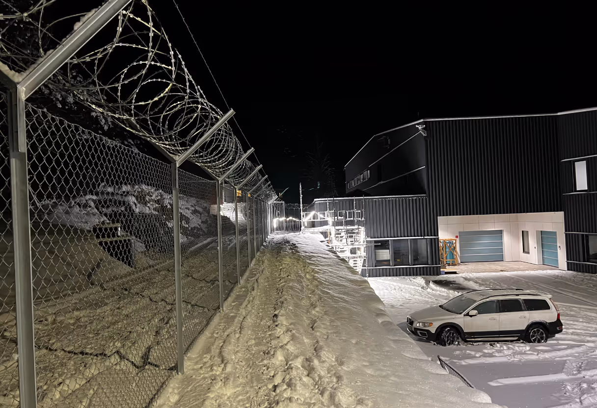 Industrielt område med piggtrådgjerde og snødekt vei ved mørk natt.