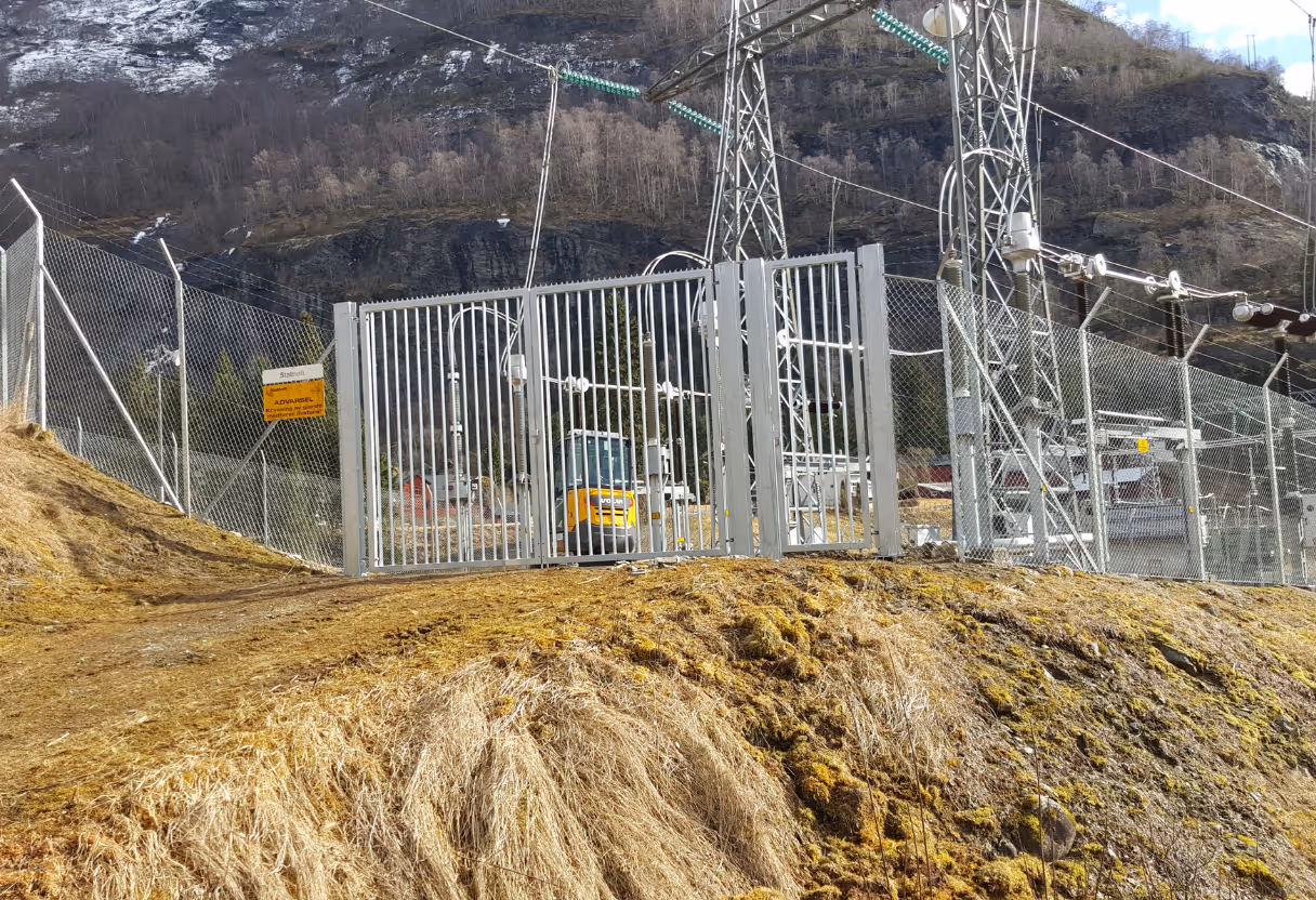 Industrielt gjerde og port rundt elektrisk transformatorstasjon i fjellområde.