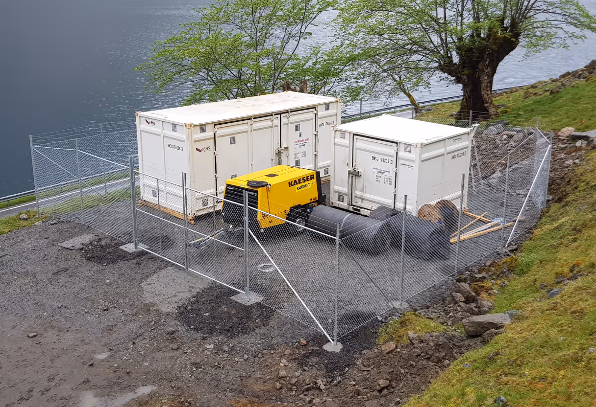 Industriområde med to hvite containere, gul maskin og sorte slanger bak sikkerhetsgjerde.