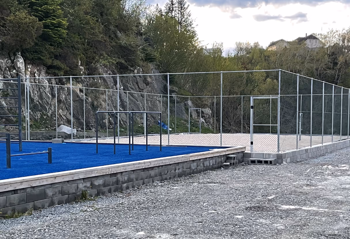Utendørs treningsområde med blå gummibelagt gulv og inngjerdet sportshall bak med nettinggjerde.