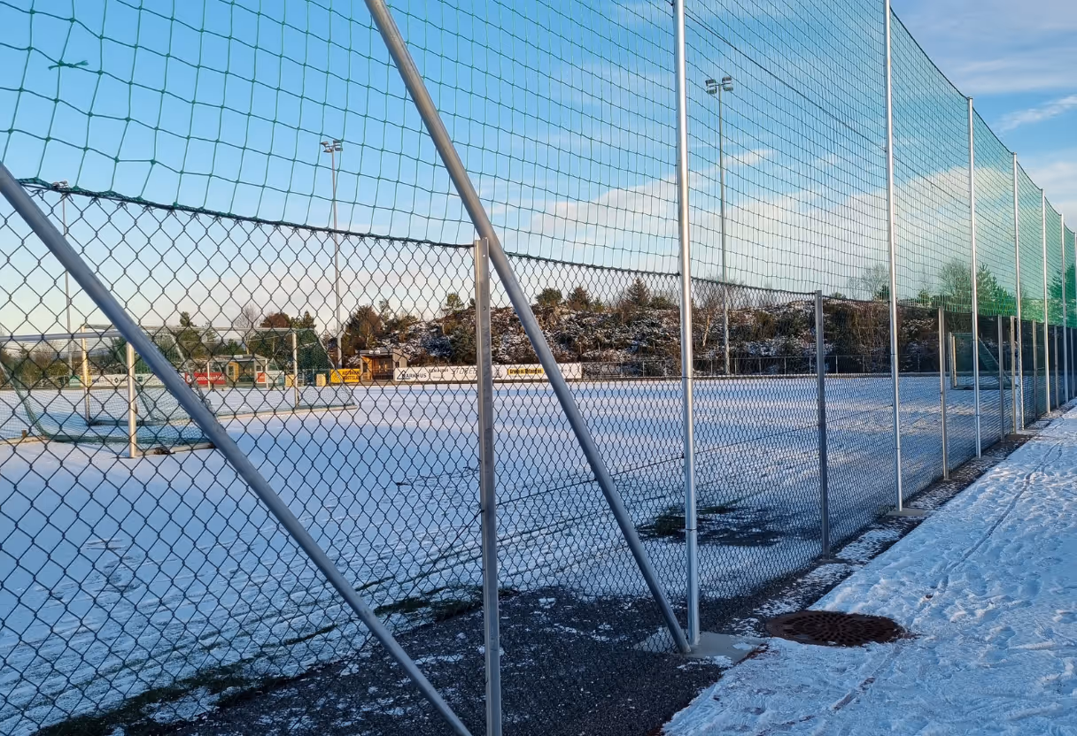 Idrettsbane dekket av snø bak et høyt gjerde med metallstolper under en blå himmel.
