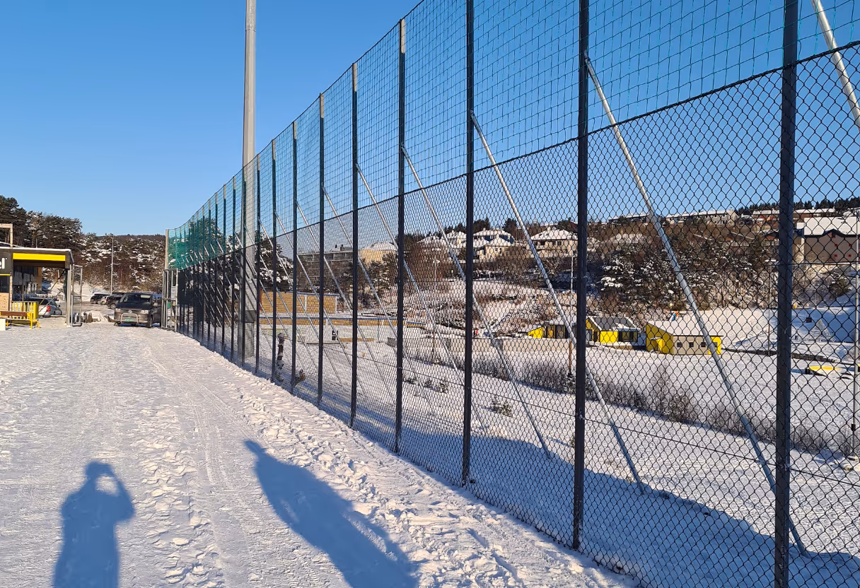 Gjerde rundt et snødekket idrettsanlegg med blå himmel og noen hus i bakgrunnen.