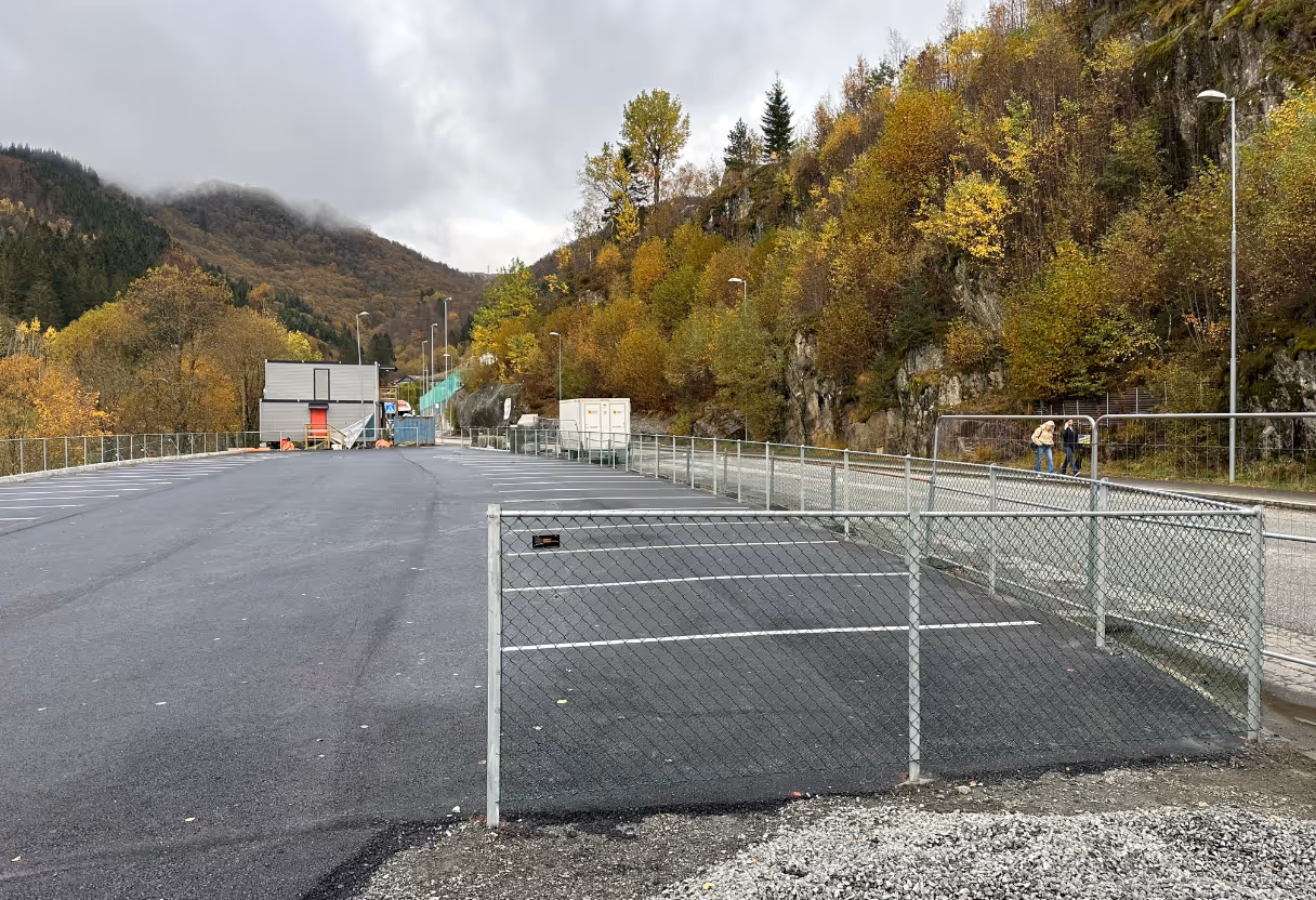 Tomt asfaltparkering med flettverksgjerde, høstfargede trær og fjell i bakgrunnen.