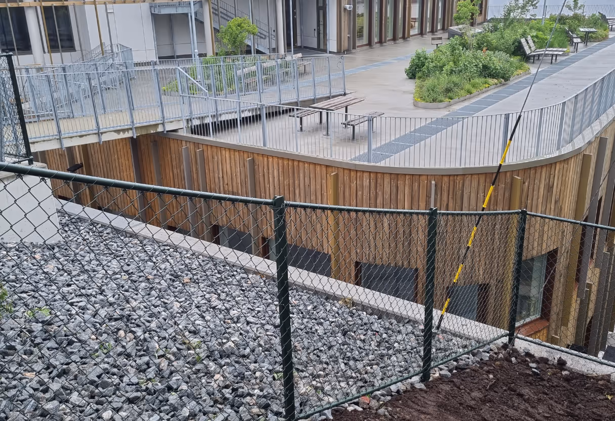Grønn metallkjettinggjerde langs skråning med grus, bak moderne bygning med benker og planter.