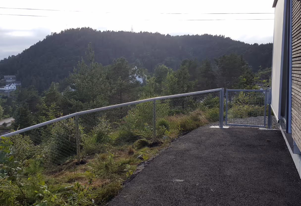 Metallgjerde med netting langs en vei ved siden av et hus og skogkledd fjell i bakgrunnen.