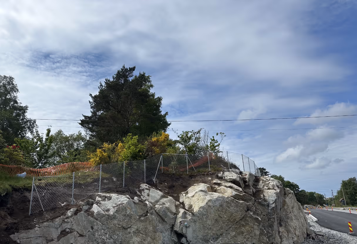 Sikring med gjerde på en steinete skrent langs en vei under en skyet himmel.